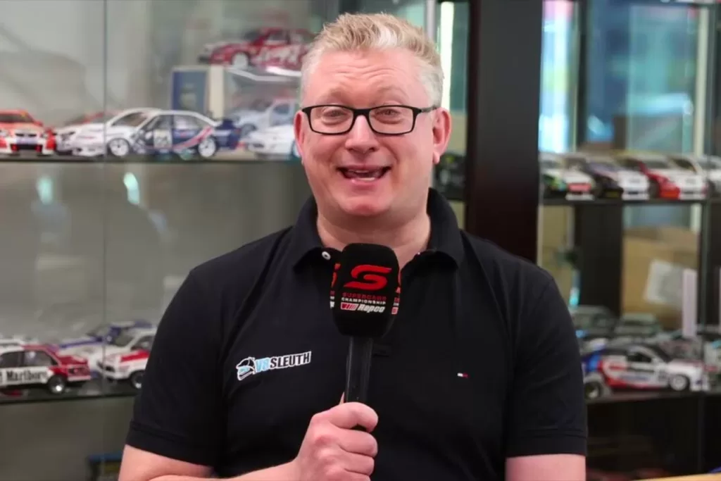 Aaron Noon with a v8 Sleuth logo holding a microphone with supercars talking to camera