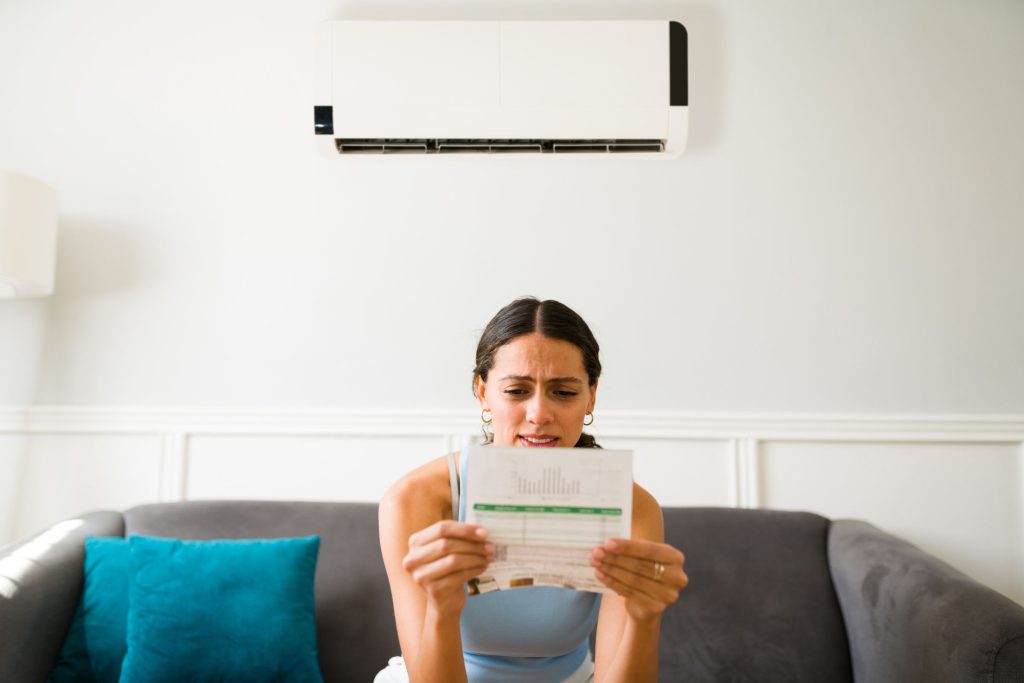 A girl sitting under air-conditioning with a real energy bill looking disappointed